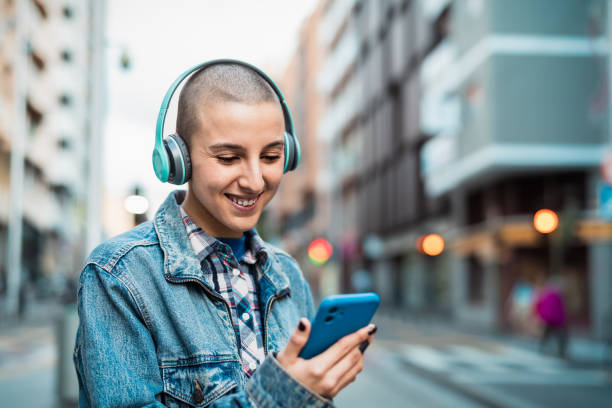 Shaved head girl using mobile smartphone and listening music while waiting for public transportation in the city street