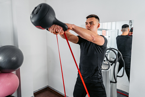 Handsome young muscular man training in the gym on a power plate with a kettlebell and a resistance band.
