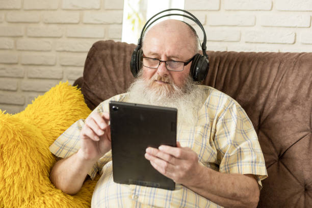istock A white senior man with a gray beard sits on a sofa at home. uses a digital tablet and wireless headphones 1480579781