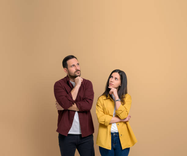 istock Uncertain young couple with hands on chins thinking for solution. Serious young man and woman dressed in shirts contemplating and looking up while standing over beige background 1480586565