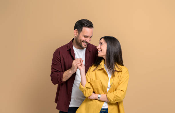 romantic young man and woman holding hands and smiling while standing over beige background. attractive couple dressed in casuals looking at each other affectionately - couple indoors studio shot horizontal imagens e fotografias de stock