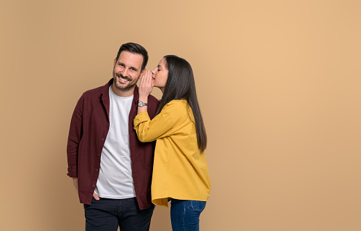 Side view of young woman covering ear of handsome smiling boyfriend while telling him gossip on isolated beige background. Couple standing together and sharing secrets with each other
