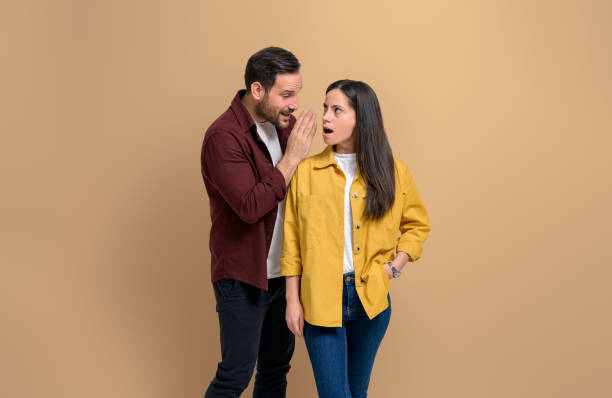 istock Young man whispering secret to shocked girlfriend looking at him with mouth open while standing isolated on beige background. Couple sharing mysterious gossips with each other 1480586871