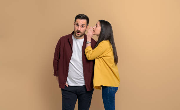 istock Side view of girlfriend whispering secret into surprised boyfriend's ear while standing isolated over beige background. Young couple sharing shocking desires with each other 1480586872