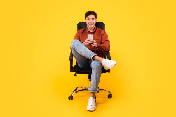 istock Happy Man Using Cellphone Sitting In Office Chair, Yellow Background 1480589609