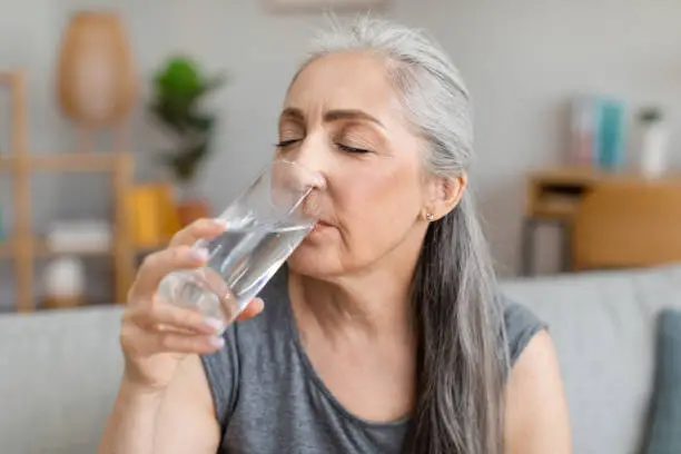 Photo of Caucasian elderly gray-haired woman drink water from glass, have useful habit in living room interior, close up