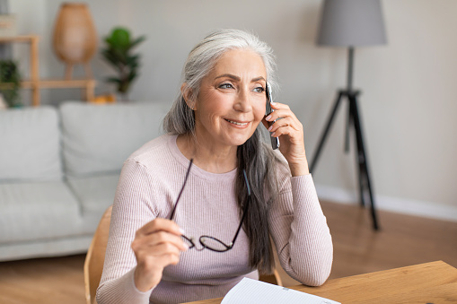 Smiling caucasian mature gray-haired lady hold glasses speaks by phone with client in living room interior, copy space. Remote work at home, communication and business during covid-19 quarantine