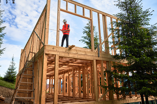 Carpenter constructing timber-framed two-story building. Man measures distances with measuring tape while dressed in workwear and hard hat. Ideology of contemporary, eco-friendly building practices.