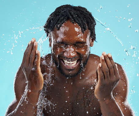 Water splash, cleaning and face with a model black man in studio on a blue background for hydration. Bathroom, skincare and hygiene with a young male washing his face for natural skin treatment