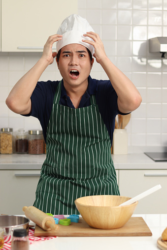 Portrait of young stressed unhappy Asian man with apron and chef hat putting his head in hands at messy kitchen, baker man chef panicked because messy cooking before preparing to bake bread.