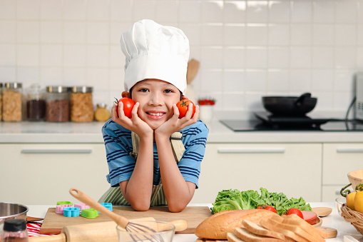 Portrait of happy smiling Asian boy kid wearing apron and chef hat, resting his chin on hands, holding and plying with red tomato during cooking healthy food at kitchen, cute child chef enjoys cooking