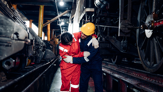 Mature foreman in workwear giving first aid to staff suffering after working injury in locomotive repair garage. Injury at work.