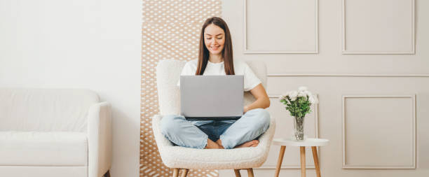 student girl using laptop at home. woman sitting on chair smiling and looking to screen young woman interior designer using laptop and sitting in lotus pose on an armchair smiling and looking to screen in white room woman sitting cross legged in chair stock pictures, royalty-free photos & images