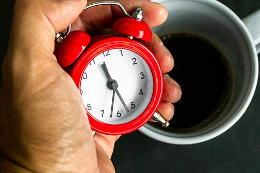 Red watch in a brown man's hand in front of a coffee mug. Mention the relationship between waking time and coffee and the neurological damage and sleep disturbances that can be caused.