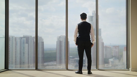 A full length portrait of a businessman standing next to windows and looking outside.