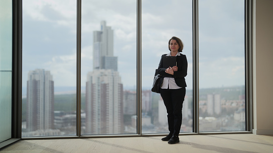 A full length portrait of a businesswoman standing next to windows and looking outside.