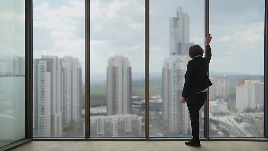 A full length portrait of a businesswoman standing next to windows and looking outside.