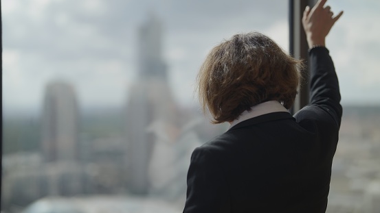 A succesful businesswoman is walking near the window and looking outside.