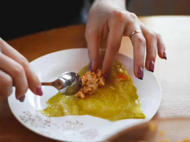 Hands of a young woman putting rice-meat stuffing on a raw sauerkraut leaf with a teaspoon in a plate, sitting at a round table in the kitchen, close-up side view. The concept of step by step instructions, home cooking, traditional recipes, national cuisine, cabbage rolls, dolma.