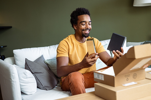 A smiling young man is opening parcels at home. He is sitting on the couch in the living room. Cardboard boxes are on the table.