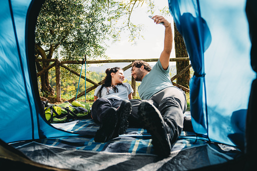 A couple is taking a selfie together near a tent at the camping. View from inside the tent. Relaxing afternoon together.