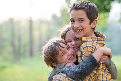 Three cute, elementary age children are looking at the camera and hugging each other playfully and lovingly, as it is comically squishing their faces together. They are outdoors in the forest in Autumn.