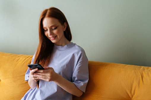 Close-up of cheerful young woman using smartphone sitting on comfy couch. Portrait of redhead female looking on mobile phone screen with smile at home. Beautiful lady relaxing chatting on cellphone.