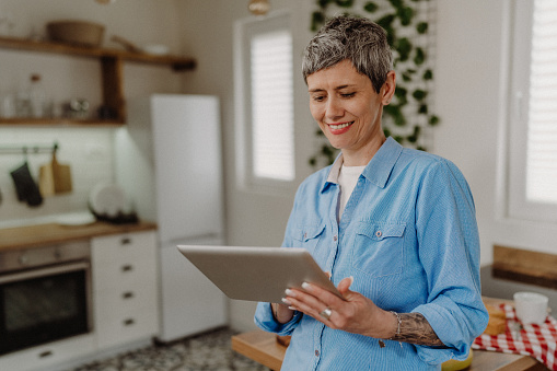 Senior woman with gray hair holding her digital tablet while standing in her kitchen