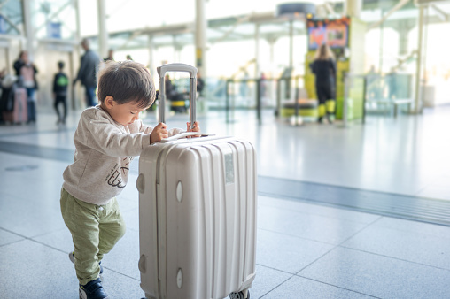 Adorable one and half year old toddler pushing a small cabin bag suitcase around the unidentified airport trying to catch his flight with his luggage
