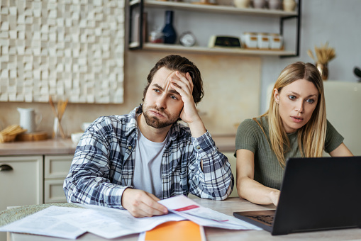 Upset european millennial man with stubble and blonde woman pay bills and taxes with laptop in light kitchen interior. Paperwork, problems with home accounting, loan and mortgage, debt due covid-19