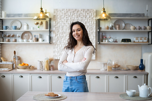 Portrait of happy young caucasian housewife posing with folded arms in kitchen interior, smiling and looking at camera while spending time at home alone, free space