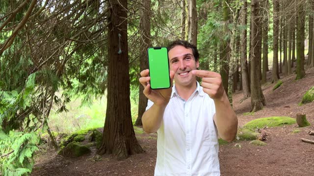 a handsome spanish man shows a phone on which a green screen chromakey he stands in a forest wearing a white shirt
