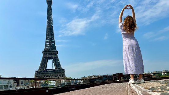 a beautiful girl in a long blue dress stands against the blue sky and the Eiffel Tower in a restaurant. She looks into the frame there is a place for advertising travel agency. High quality photo