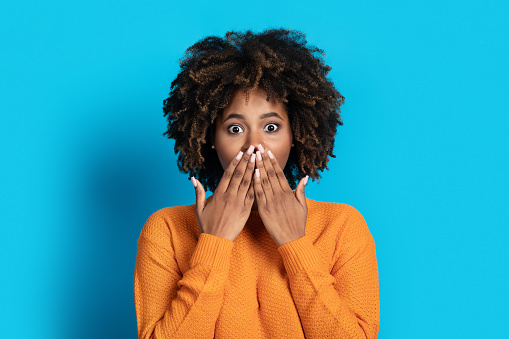 Surprised emotional cute attractive young black woman with bushy hair covering mouth, looking at camera, isolated on blue studio background. Excitement, shock human emotions concept