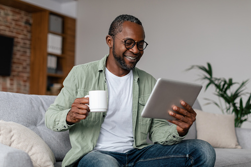 Smiling mature african american man in glasses drinking coffee from cup and watching video on tablet in living room interior. Chat in social networks remotely, new normal, app for working at home