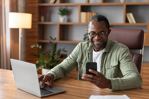 Glad busy handsome adult black businessman in glasses with computer typing on smartphone in home office interior. App for business, gadget for work, data analysis and customer support, covid-19 virus