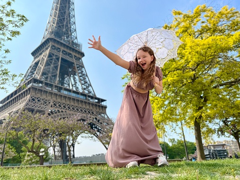 a cheerful teen girl stands against the backdrop of the Eiffel Tower in a beautiful dress, she raised her hand, you can advertise a travel agency trip to Paris France. High quality photo