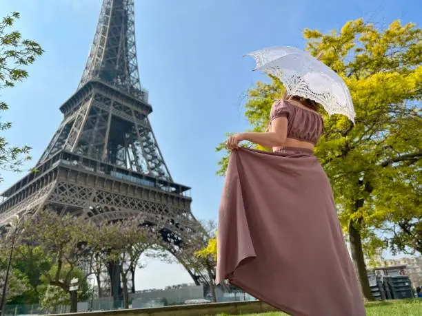 Photo of from the back we see a girl in a beautiful long brown dress in a retro style with an umbrella who walks towards the eiffel tower holding the dress with one hand