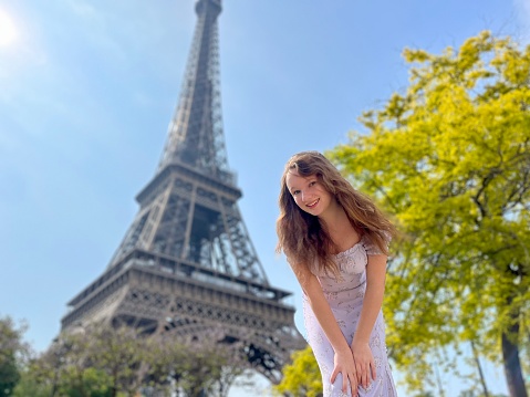 a beautiful teenage girl looks into the frame leaning against the backdrop of the Eiffel Tower She smiles and seems to be showing come here great advertisement for a trip to Paris. High quality photo