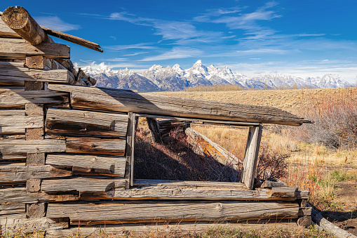 Remains of an abandoned homestead cabin overlooking the snow coveredTeton Mountain Range in Grand Teton National Park Wyoming with blue sky and clouds .