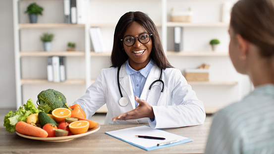 Friendly nutritionist black woman giving consultation to patient and demonstrating plate with fresh fruit and vegetables, sitting at workplace in clinic. Nutrition and diet concept