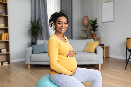 Cheerful young black pregnant lady future mom with big belly do exercises on fit ball, feels baby movements in living room interior. Expecting child, sports training, body and health care at home