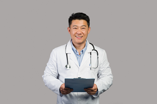 Smiling mature chinese man therapist in white coat with stethoscope use tablet, isolated on gray background, studio. Doctor recipe for treatment, medical examination, health care and support in clinic