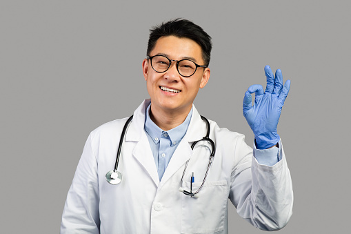 Happy middle aged asian male therapist in white coat, protective gloves with stethoscope showing ok sign, isolated on gray background. Doctor recommendation and advice for health care and medicine