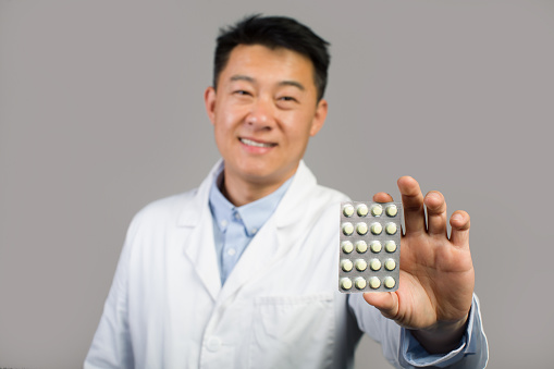 Smiling middle aged chinese male therapist in white coat showing pills, recommended medical treatment, isolated on gray background, studio, close up. Pharmacy health care, doctor advice, offer and ad