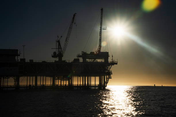 An offshore oil rig drilling and fracking operation, illuminated by bright lights, operates on calm seas at night. At dusk, the oil platform can be seen near Los Angeles, California.