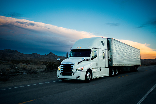 White semi-truck traverses a 2-lane highway in the Arizona desert at dusk, its imposing frame set against a dramatic cloudscape, as it journeys through the rugged terrain