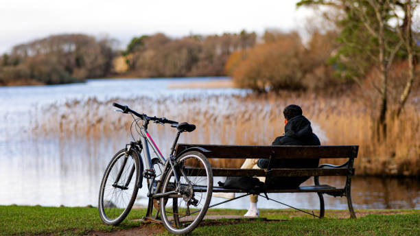 solo junto al lago joven con una bicicleta se sienta en el banco y fuma, sentado en el banco contra la vista de la naturaleza, relajándose en la naturaleza, aburrido de que todo se aleje de la ciudad, descansando su alma en la naturaleza cuando está lejo - bench mountain park sitting fotografías e imágenes de stock