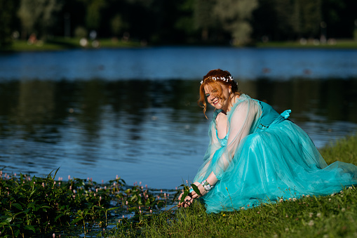 Portrait of redhead young bride in blue dress sitting by pond in park. Wedding or prom photo.