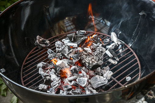 Mobile round grill with glowing charcoal, small flames and smoke, preparation for a barbecue party, copy space, selected focus, narrow depth of field